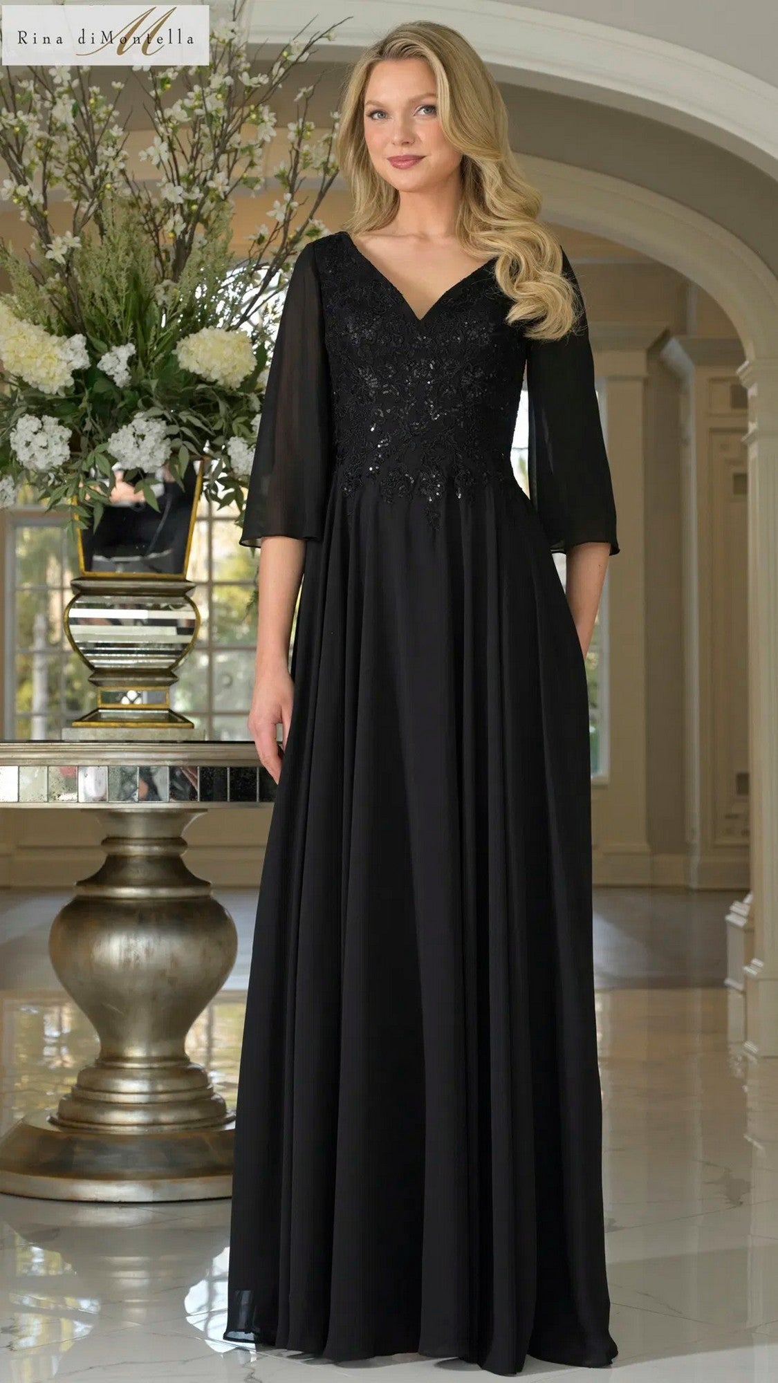 A woman stands indoors in the Rina Di Montella RD3241, a long black formal dress featuring a floral-embroidered bodice, three-quarter sleeves, and an elegant flowing A-line skirt. She poses near a large floral arrangement and ornate table.
